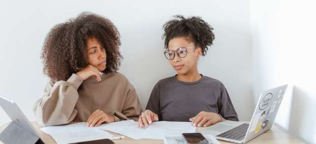 Zwei Studentinnen sitzen mit Laptop und Unterlagen am Tisch und rechnen die Studiengebühren durch.