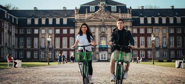 Zwei Studierende mit Hochschulzugangsberechtigung radeln vor dem Schloss ihrer Universität.