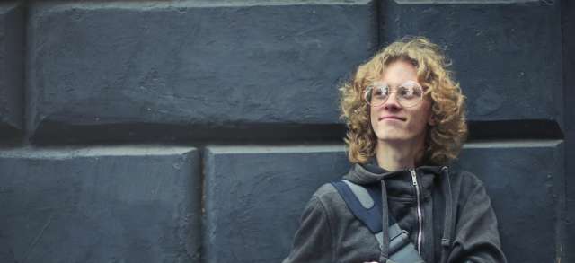 Ein junger Student mit Büchern im Arm lehnt an einer dunklen Wand und blickt zuversichtlich in Richtung Stipendium.