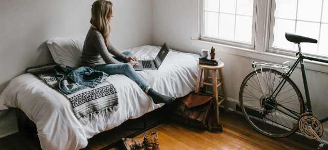 Eine Studentin sitzt mit ihrem Laptop auf dem Bett ihres schlicht eingerichteten Zimmers im Studentenwohnheim.