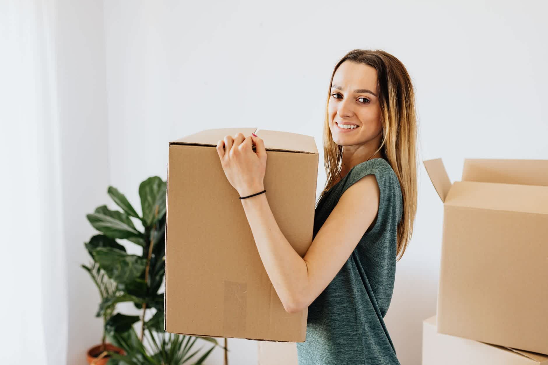 Junge Frau trägt bei der Wohnungssuche einen Umzugskarton in ihre neue Wohnung.