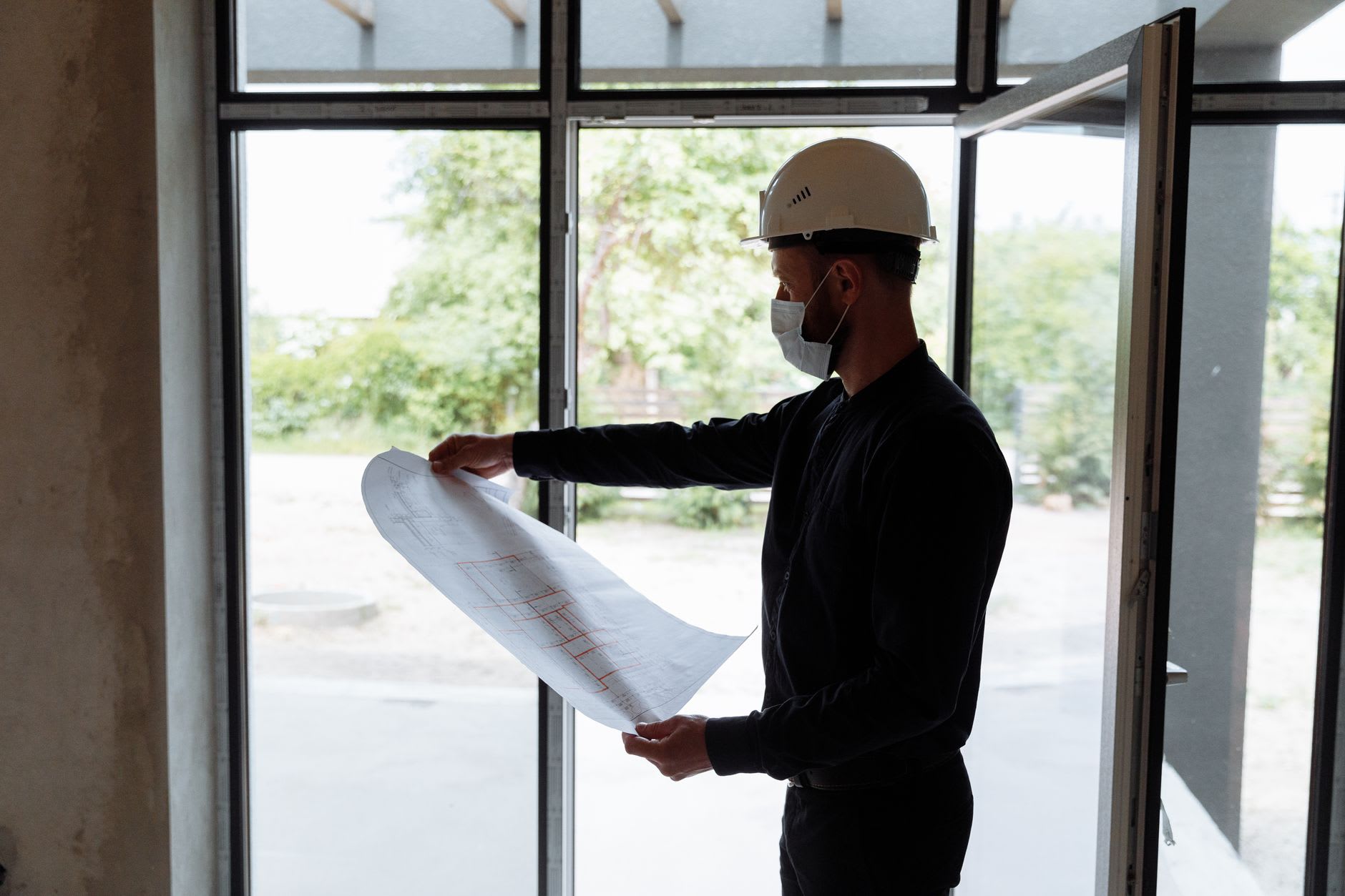 Bauingenieur mit Schutzhelm und Maske studiert am Fenster einen Bauplan.