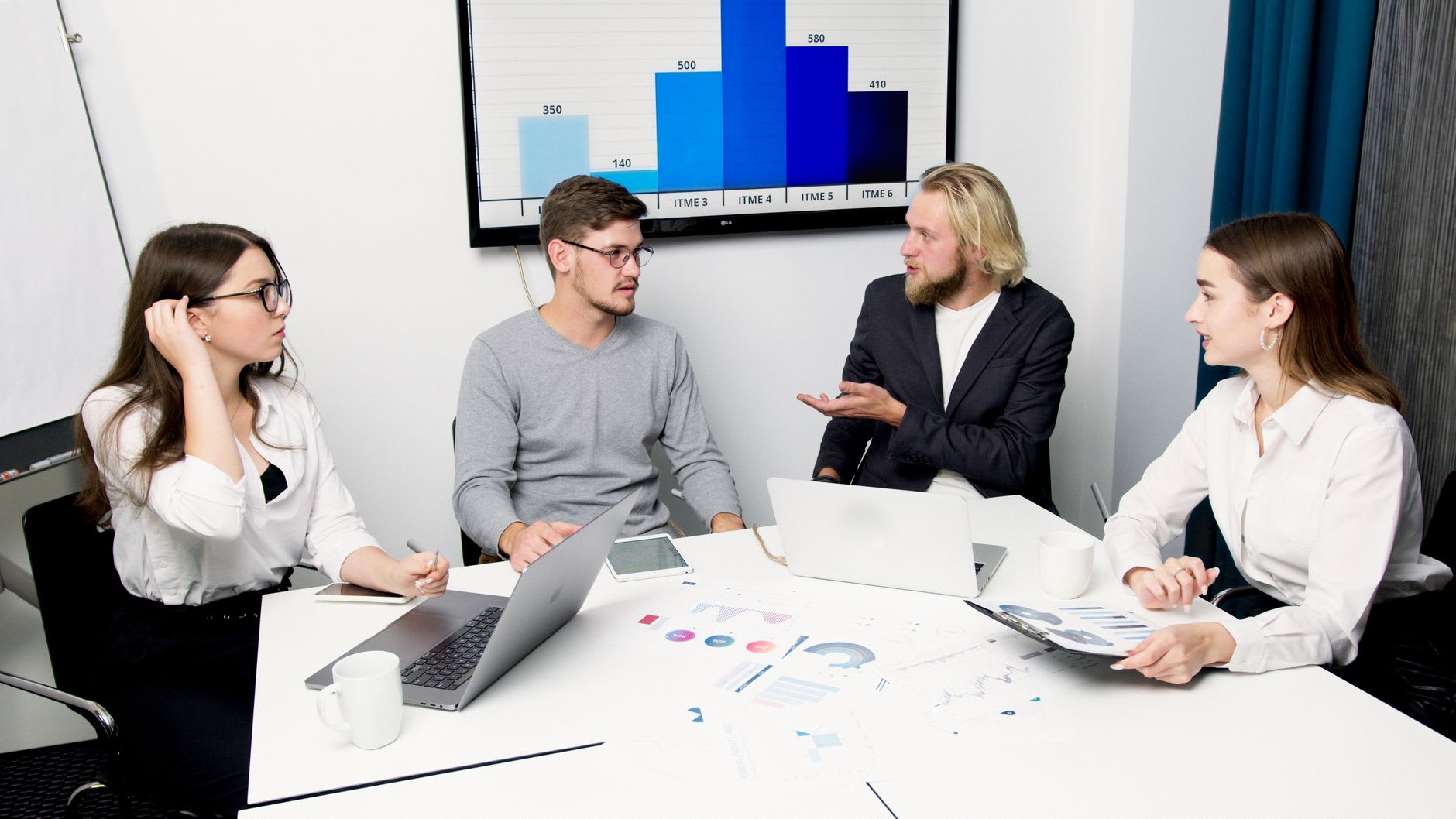 Vier junge Berufstätige besprechen im Meeting vor einem Balkendiagramm betriebswirtschaftliche Kennzahlen.