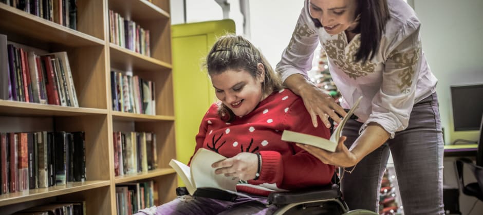 Young woman using a wheelchair in the library with another woman who is standing and looking at books with her.