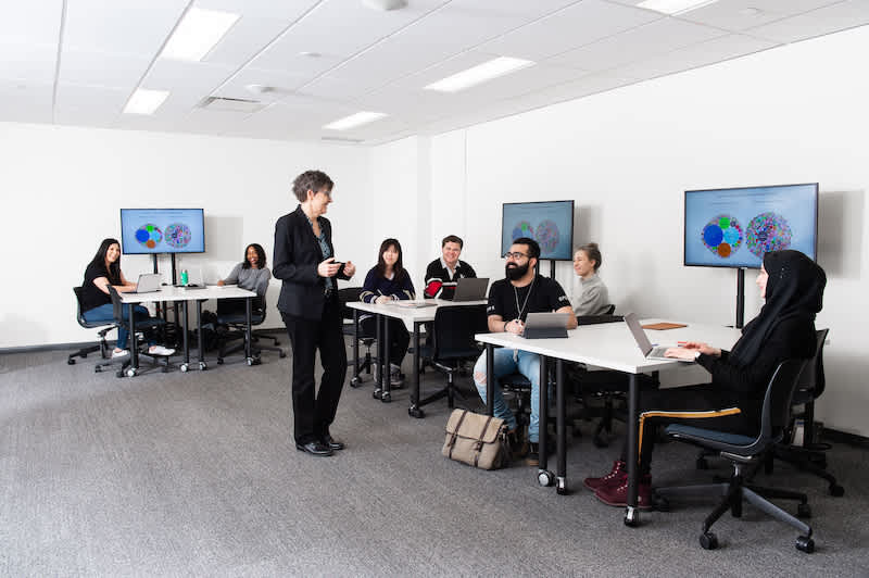 Profesor de pie en el aula de alta tecnología, con estudiantes en mesas con pantallas de computadora