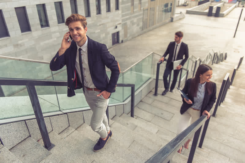 Beautiful young business people in classic clothes are coming upstairs in the office building. Guy in the foreground is talking on the mobile phone and smiling