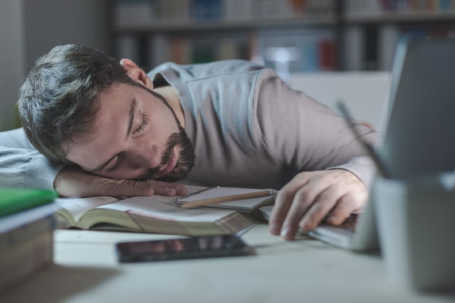 Young exhausted man in the office late at night sleeping on his desk on a book, restlessness and overwork concept