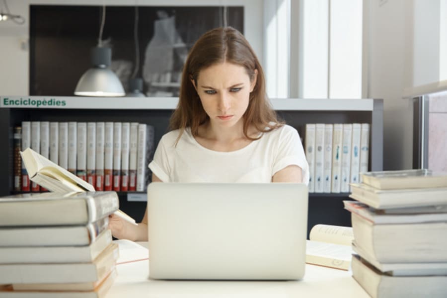 Portrait of female student sitting at the bookshelf and using laptop in the library, reading books, writing thesis with serious face expression. Young Caucasian woman working on her home assignment