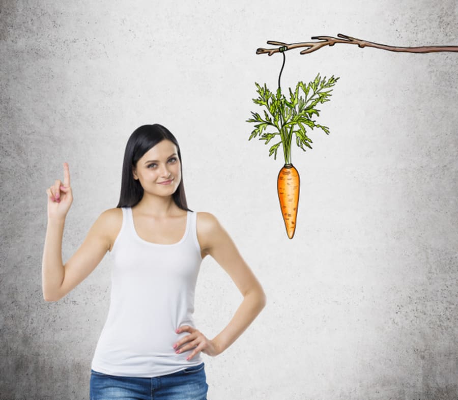 A young pretty woman with an index finger up standing next to a picture of a carrot tied to a branch. Concrete background. Concept of a new idea and future praise.