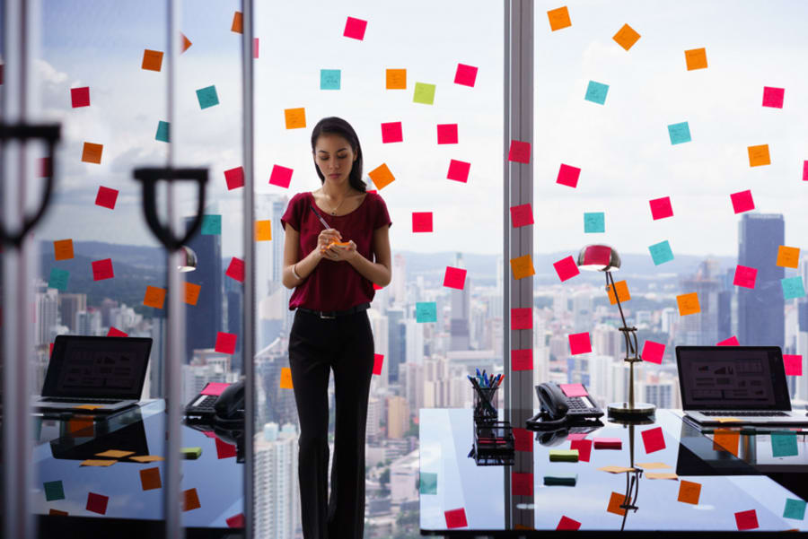 Mixed race secretary working in modern office in skyscraper, writing and sticking adhesive notes with tasks on window.