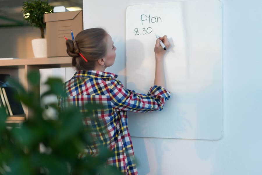 Young woman writing day plan on white board, holding marker in right hand. Student planning schedule rear view portrait.