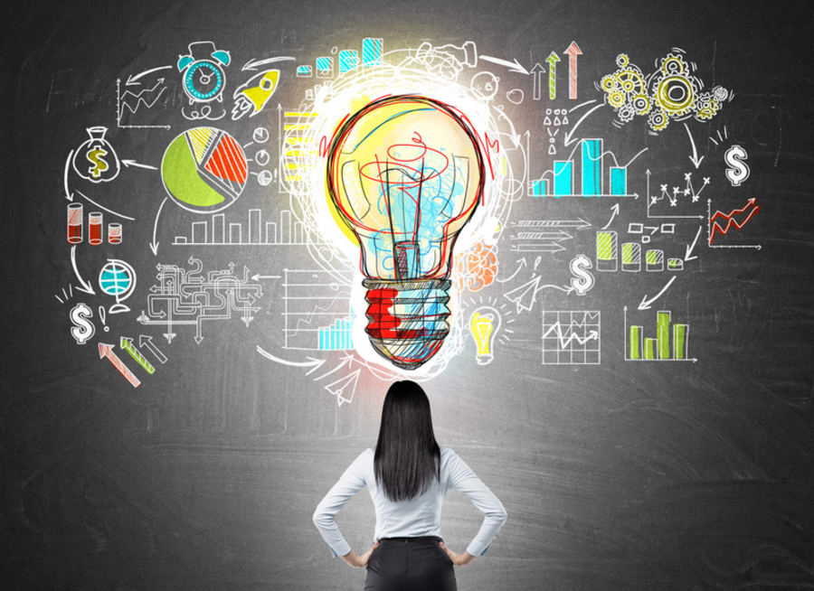 Rear view of businesswoman with hands on waist looking at colorful startup sketch on chalkboard with large light bulb icon in middle. Concept of bright idea.