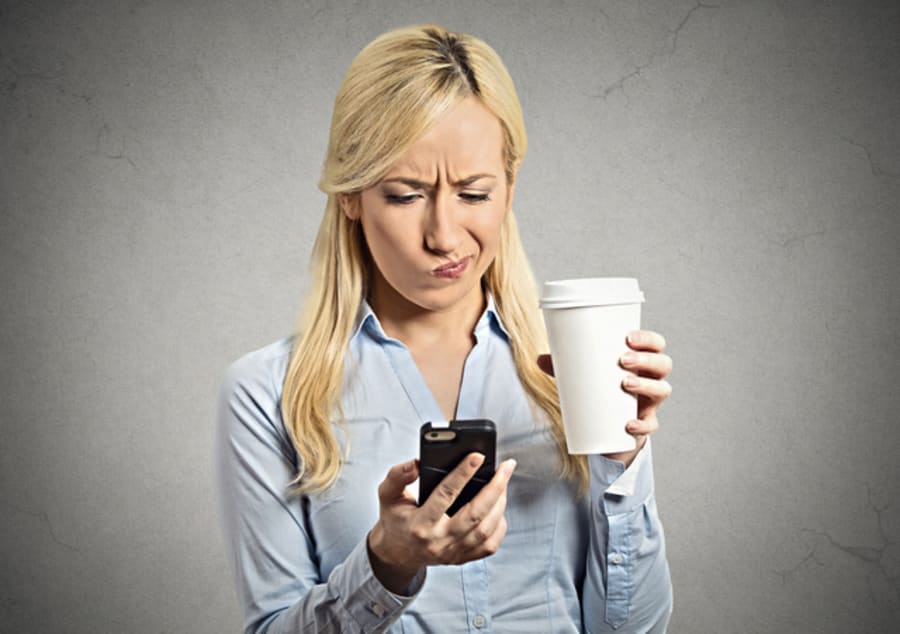Closeup portrait serious worried business woman reading bad news on smart phone holding mobile drinking cup of coffee isolated grey wall background. Human face expression, corporate executive emotions