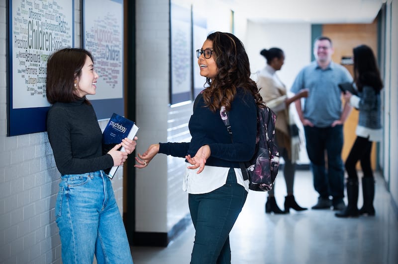 Dos mujeres jóvenes hablando en el pasillo - estudiantes y estudiantes en el fondo