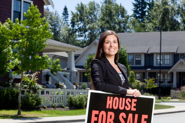 Professionally dressed real estate woman outside in a neighborhood leaning on a for sale sign.