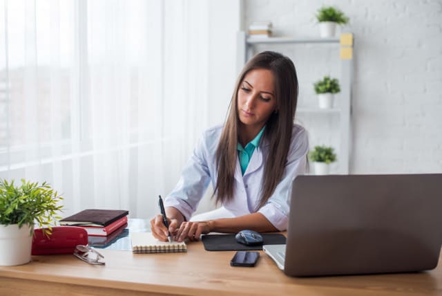 Portrait of physician doctor working in medical office workplace writing prescription sitting at desk.