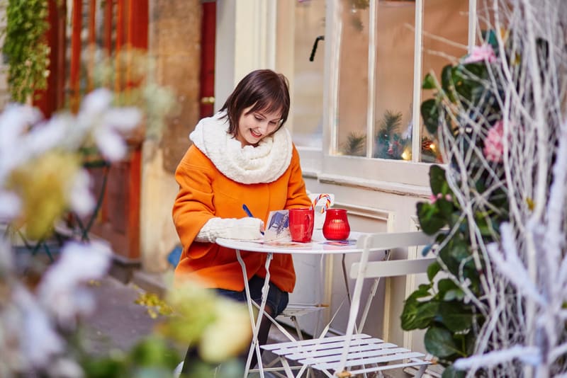 Cheerful young girl writing Christmas cards or New Year resolutions in Parisian outdoor cafe