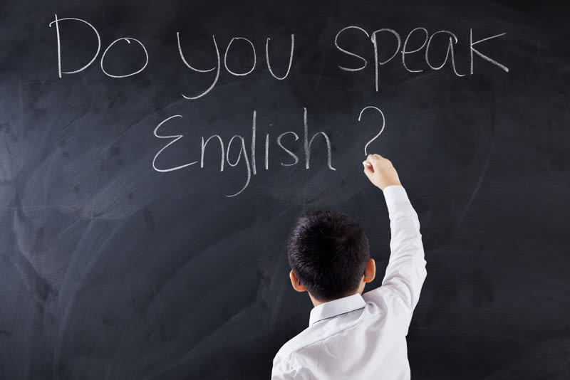 Male primary school student writes text of Do You Speak English on the blackboard