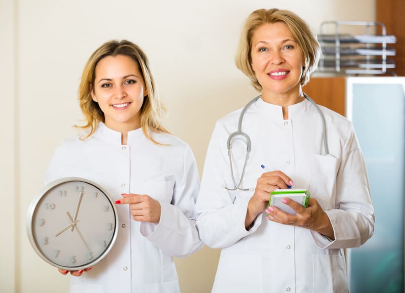 Two smiling female doctors in white overalls holding clock in modern clinic