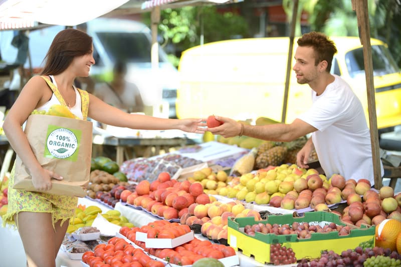 Small business owner at an open street market, handing out a fruit to a consumer carrying a shopping paper bag with a 100% organic certified label full of fruit and vegetables.