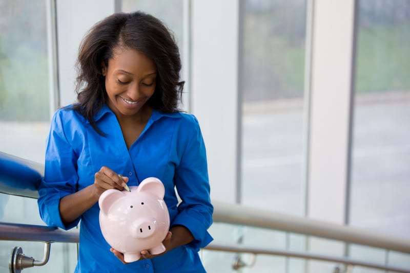 Closeup portrait joyful, smiling businesswoman in blue shirt, holding pink piggy bank, isolated indoors office window and rail background. Financial budget savings, smart investment concept