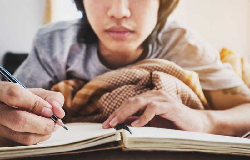 Man writing with pencil on notebook on bed in morning