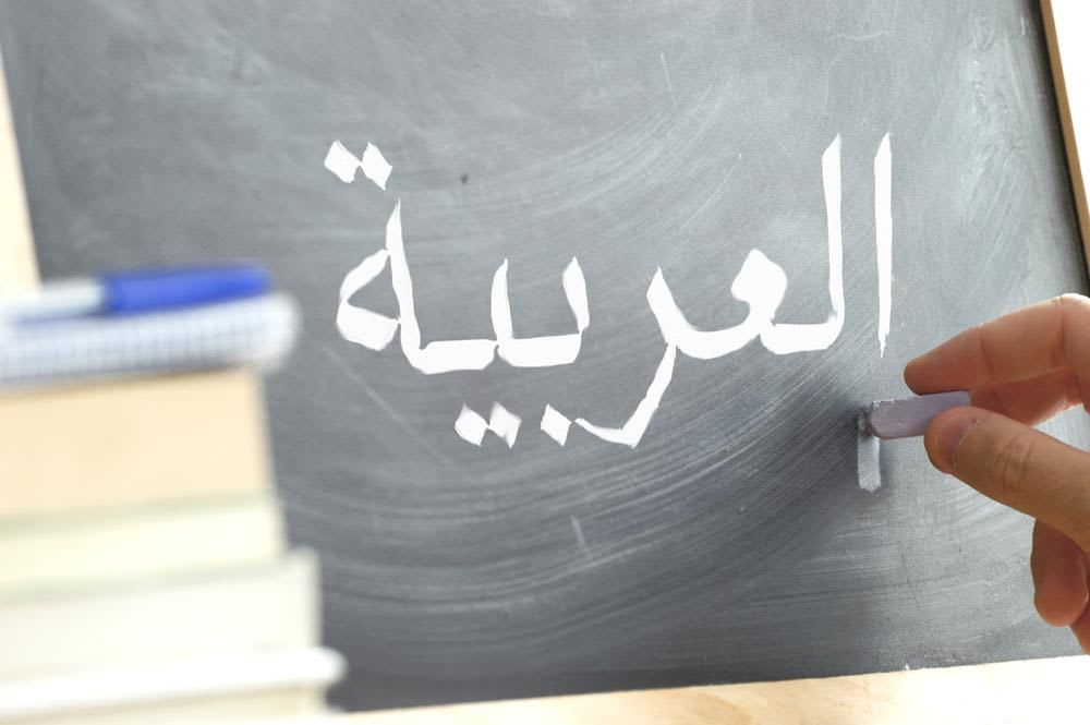 Hand writing on a blackboard in a language class with the text "Arabic" written on it. Some books and school materials.
