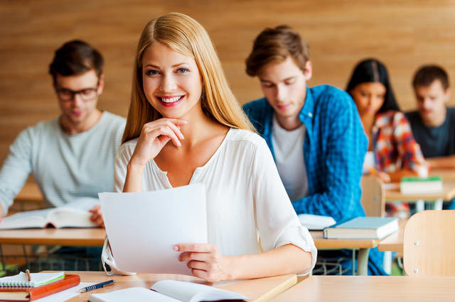 Working toward A. Smiling young woman holding hand on chin and looking at camera while sitting at front desk in the classroom