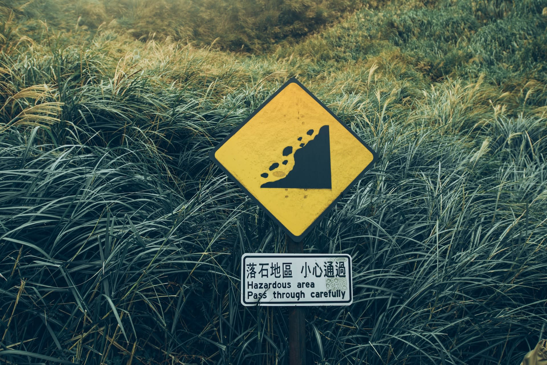 Hazard sign in a grassy field warning of falling rocks in English and Japanese