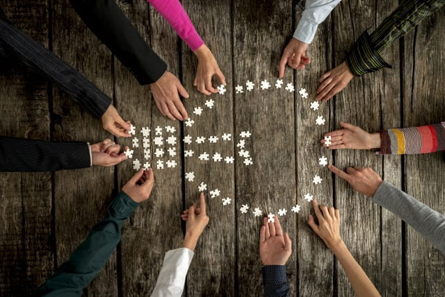 Teamwork and cooperation concept - group of twelve people, male and female, assembling a light bulb shape with blank puzzle pieces on a rustic textured desk, top view.