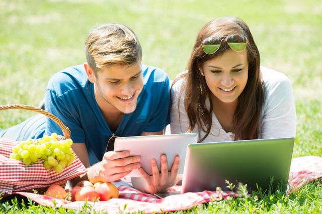 Happy Couple Using Digital Tablet And Laptop While Lying In Park