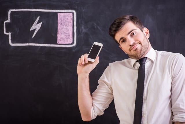 Young man is holding  his smartphone with battery low while standing against the blackboard with drawing buttery low