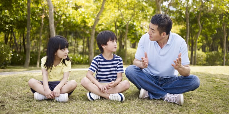 asian father and two children sitting on grass having an interesting conversation, outdoors in a park.