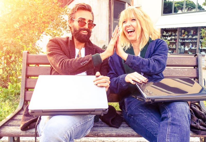 Creative business couple hands up doing high five outdoor -  Cheerful hipsters holding pc laptop and gesturing in a city park - Concept of teamwork and fun together at sunset light with vintage filter