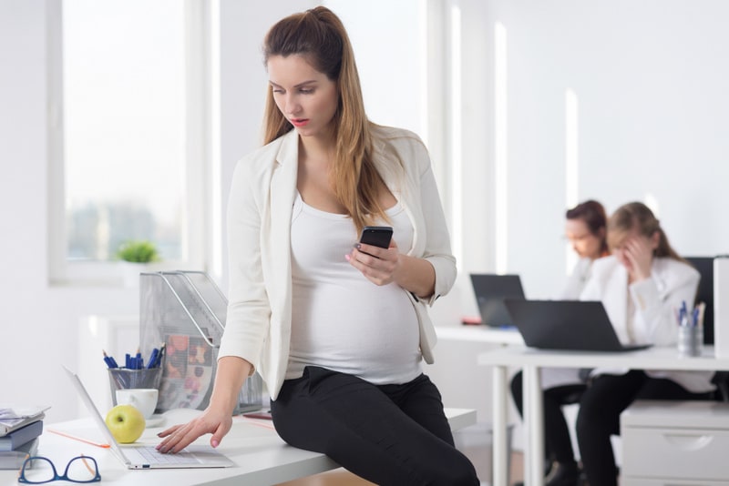 Pregnant boss sitting on a desk in a light office, holding a cellphone