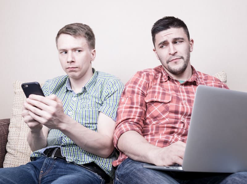 Two young guys operating with gadgets sitting apart on sofa
