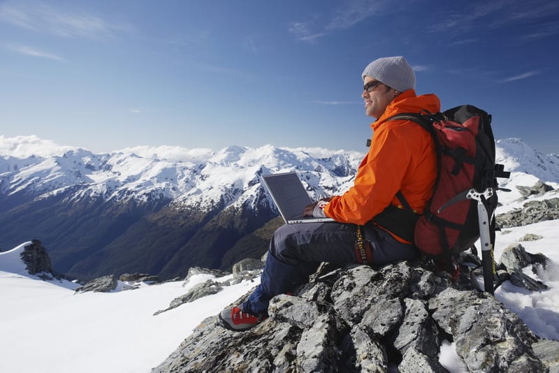 Side view of a male mountain climber using laptop on mountain peak