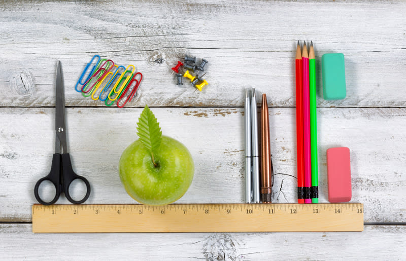 Green apple, pens, pencils, erasers, paper clips, tacks and scissors lined up with ruler on top of white desktop. Education concept.