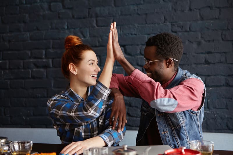 Teamwork, cooperation, interracial friendship and partnership concept. Cheerful young African male in trendy clothes and glasses giving high five to redhead pretty girl while celebrating good news