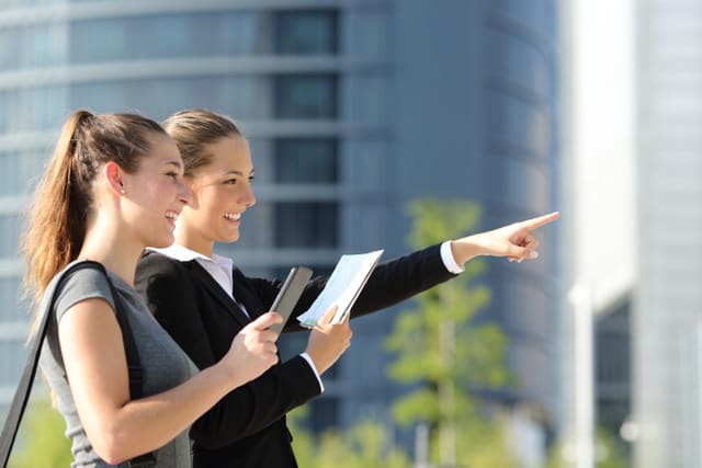 Two businesswomen searching location with mobile phone gps and paper map with office buildings in the background