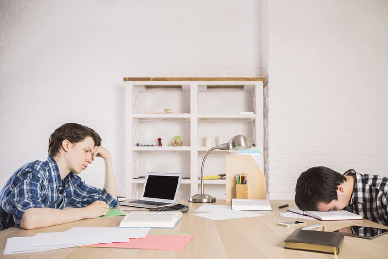Two college students tired of studying for exam at large wooden table with blank laptop, homework, supplies and other items on white brick wall background. Mock up