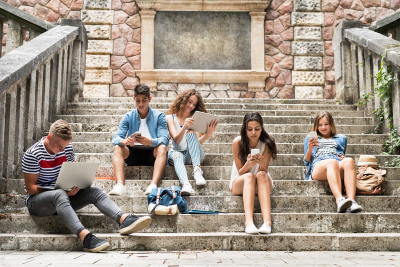 Teenage students with gadgets outside on stone steps.