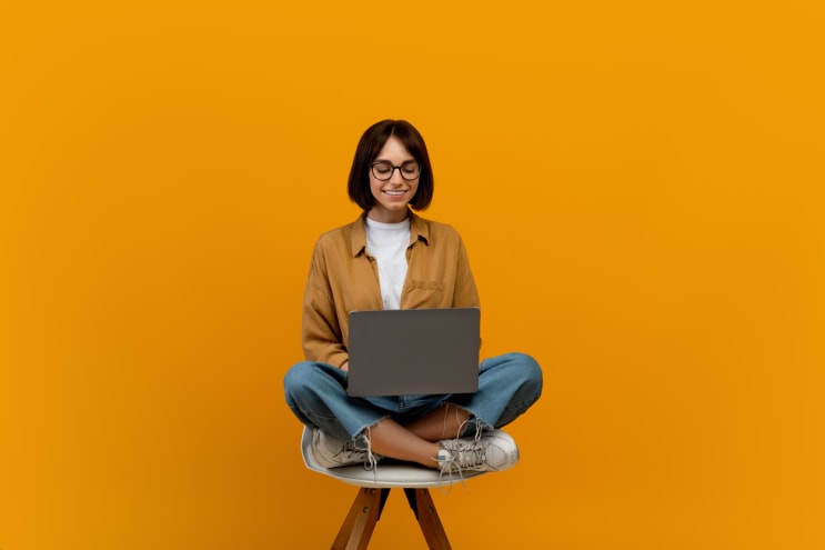 E-learning concept. Female student using laptop computer while sitting in chair over yellow studio background, copy space. Online lectures and courses, educational webinar