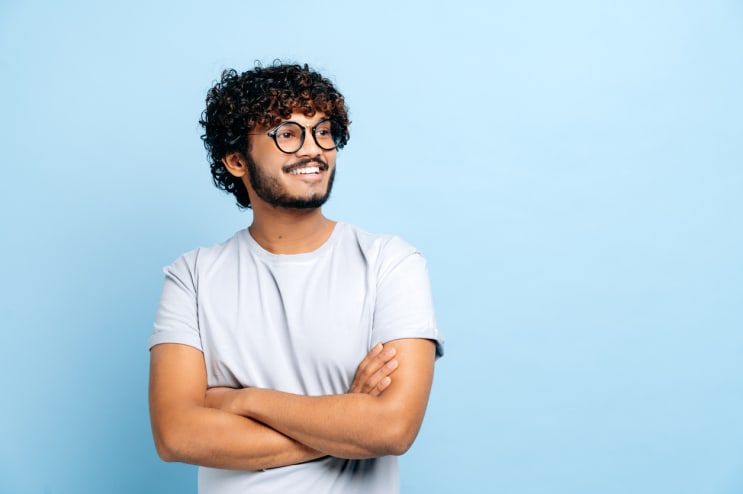 Attractive positive Indian or Arabic curly haired guy with glasses, wearing a basic t-shirt, student or freelancer, standing over isolated blue background, with arms crossed, looks to the side, smiles stock photo